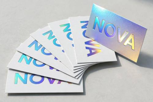 A fanned stack of business cards with the wordmark 'NOVA' foil-stamped large across each card, one card standing upright catching the light. Overhead three-quarter view on a pale gray table. Rainbow foil on pure white. Sleek and premium. Holographic foil product shot: iridescent rainbow foil that shifts from cyan to violet to gold across its surface, crisp foil-stamped edges, everything else printed on matte bright white or pale gray stock, arranged on a clean light surface with soft shadows, sharp focus. Premium and futuristic.