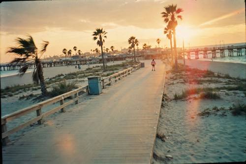 A roller skater gliding along a beach boardwalk at golden hour, 1980s, headphones and striped knee socks, palm trees and a pier behind. Wide shot with the skater small in the frame, horizon slightly tilted. Warm low sun with a soft lens flare. Peach, faded turquoise and sand. Breezy and joyful. A real snapshot scanned from 35mm color negative film: coarse grain across the whole image, faded warm colors with a slight green cast in the shadows, milky lifted blacks, a faint red glow around bright lights, soft focus, amateur framing. Not sharp, not glossy, not HDR, not a modern digital photo.