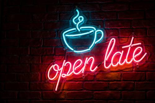 A neon sign reading 'open late' in pink script with a cyan coffee cup icon, mounted on dark brick. Straight on, slightly off-center. Hot pink and cyan on dark red brick. Warm and inviting. Neon sign photo: glowing glass neon tube lettering and simple line icons, bright saturated glow with soft halos and light spilling onto the surface behind, hot pink, electric cyan and orange, visible tube bends and small mounting clips, low ambient light. A real photograph, moody and electric.