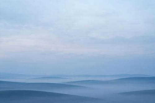 A desktop wallpaper of misty rolling hills in layered blue tones at dawn, a soft gradient sky taking up most of the frame, minimal and calm.